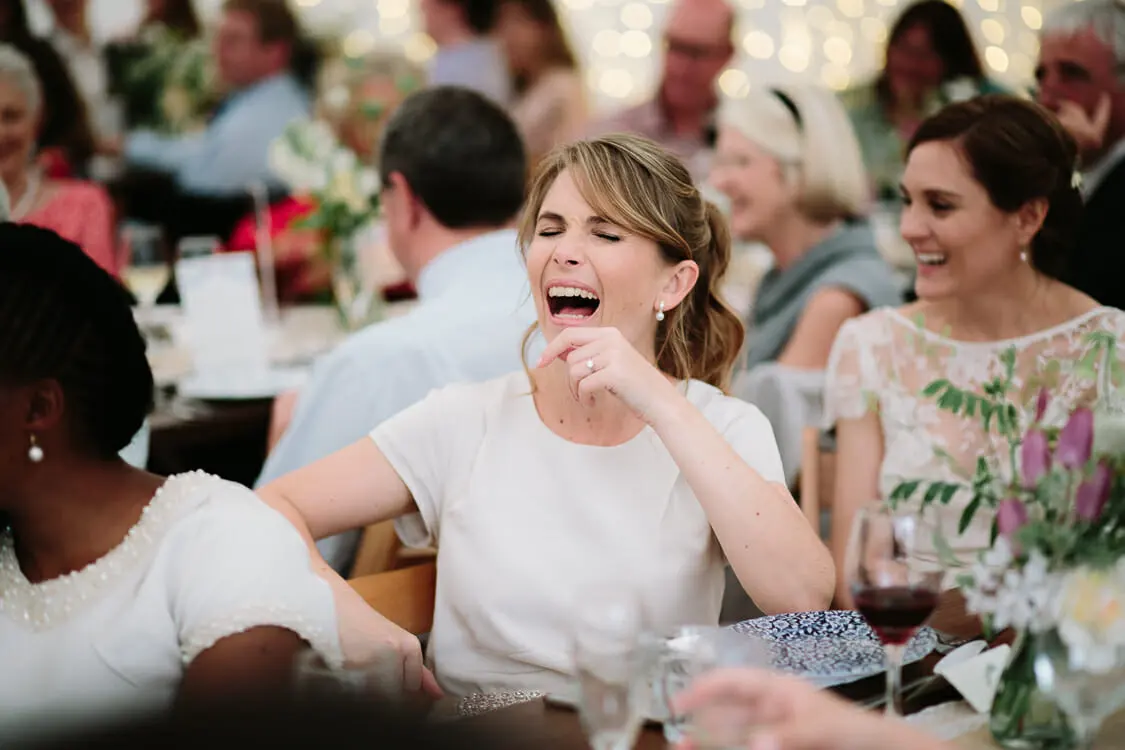 Guest in white laughing with her eyes shut at a reception table during the speeches