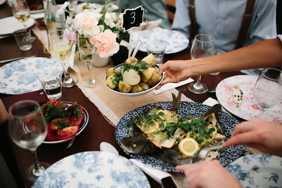 Guests passing dishes of new potatoes, baked fish and tomato salad along a family-style table