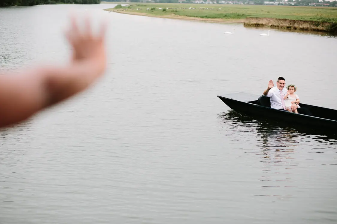 Guest and a small girl in a flower crown waving from a punt on the river, with swans behind