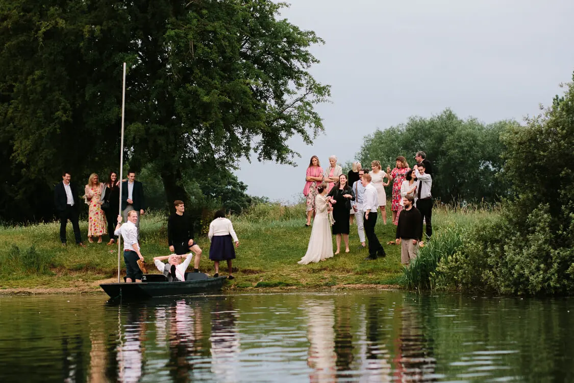 Wedding guests and the bride on a riverbank as a guest punts a boat on the river at the Perch