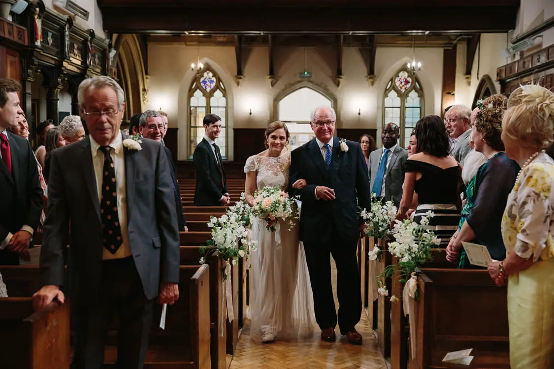 Bride walking down the chapel aisle on an older man's arm as standing guests look on