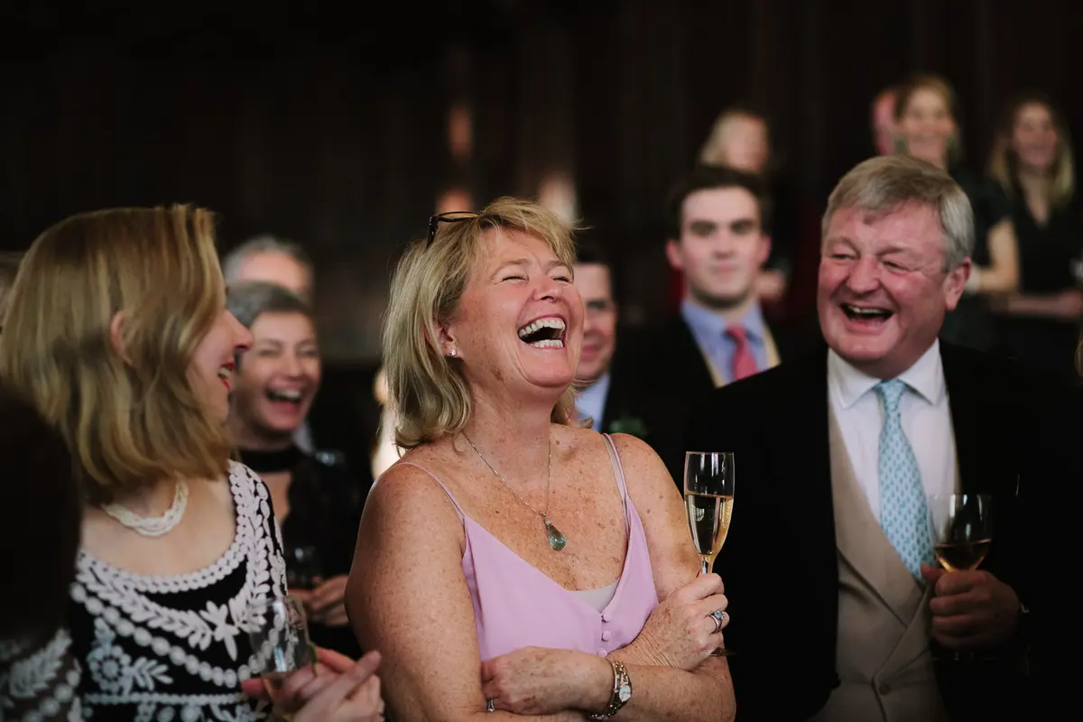 Guests holding champagne glasses roaring with laughter during the wedding speeches