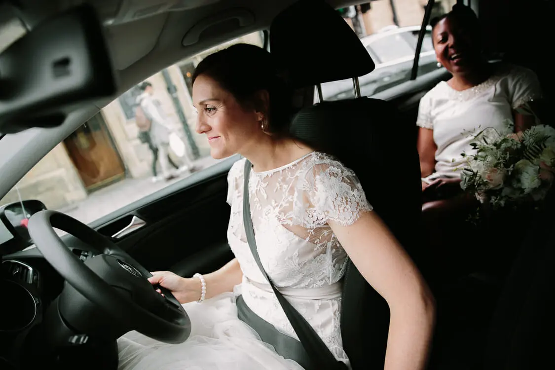 Smiling bride in lace at the wheel of a car as a friend holding the bouquet laughs in the back