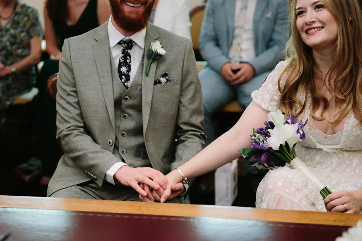 Bride and groom holding hands at the registrar's table, the bride holding purple and white flowers