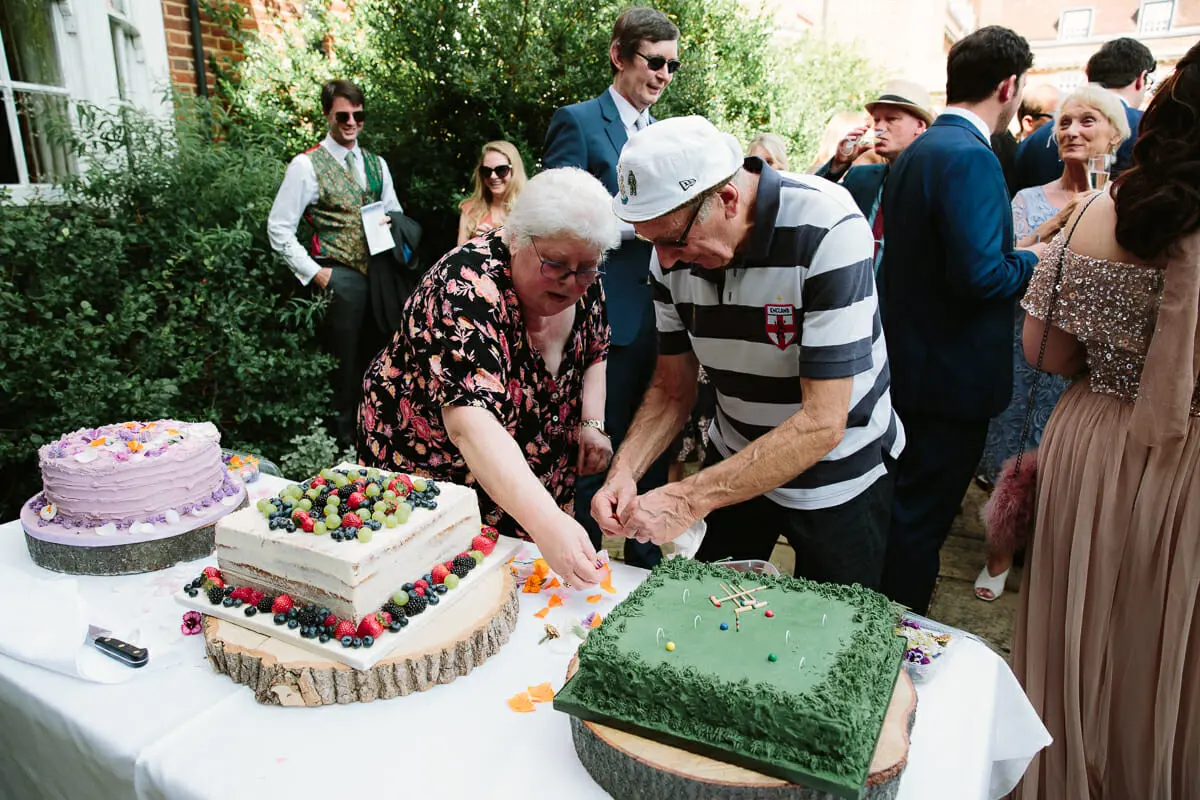 Older couple cutting a wedding cake topped with berries beside a green cricket-pitch cake at an outdoor reception