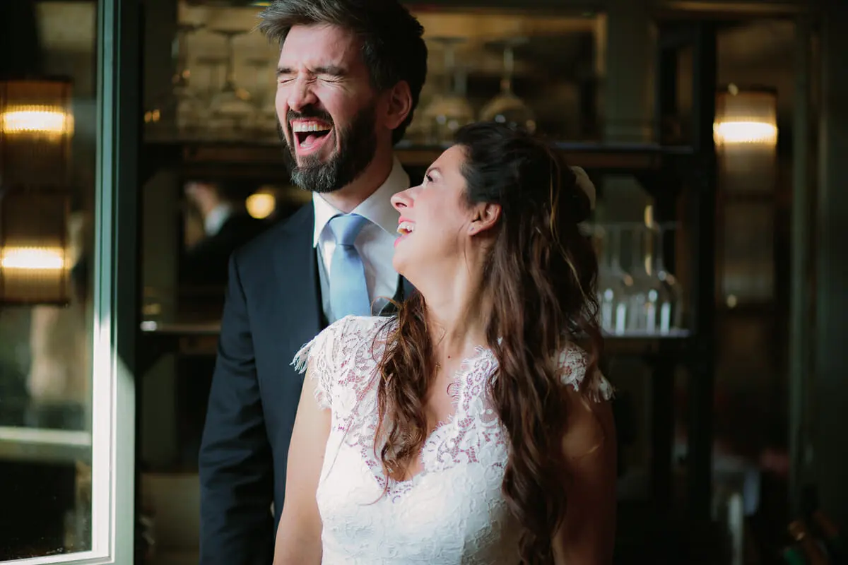 Bride and groom laughing together in warm light by a window in a bar