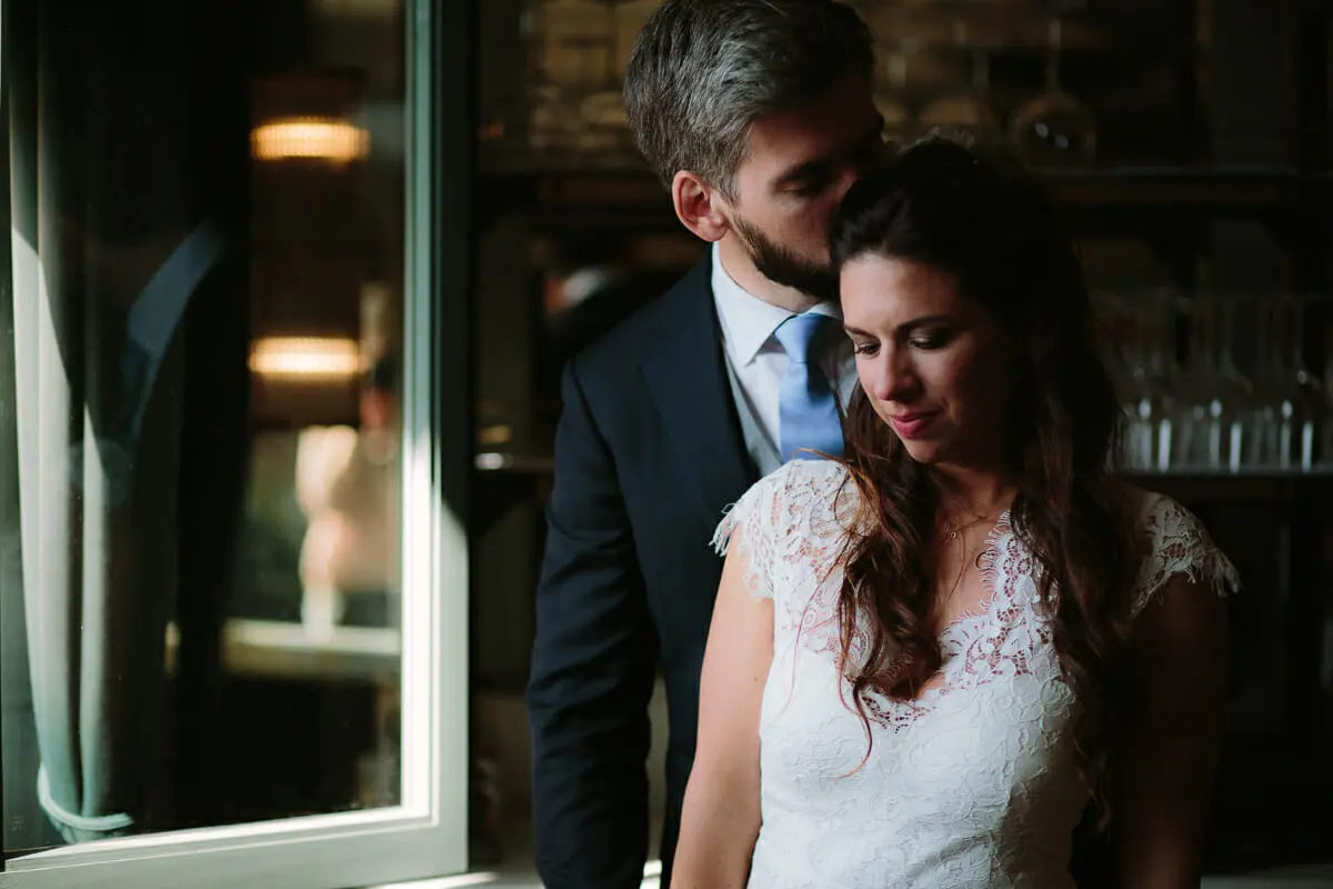 Groom kissing the top of the bride's head beside a window in soft light