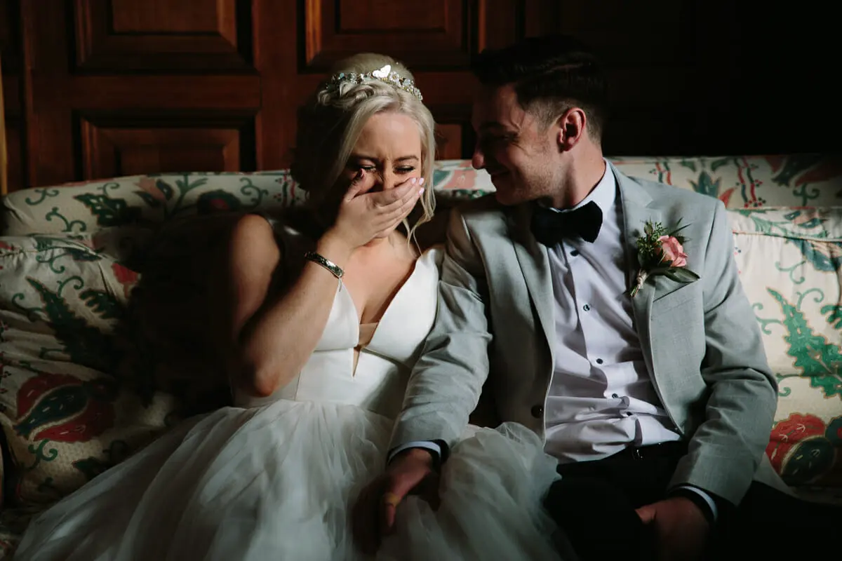 Bride covering her face as she laughs beside the groom on a floral sofa