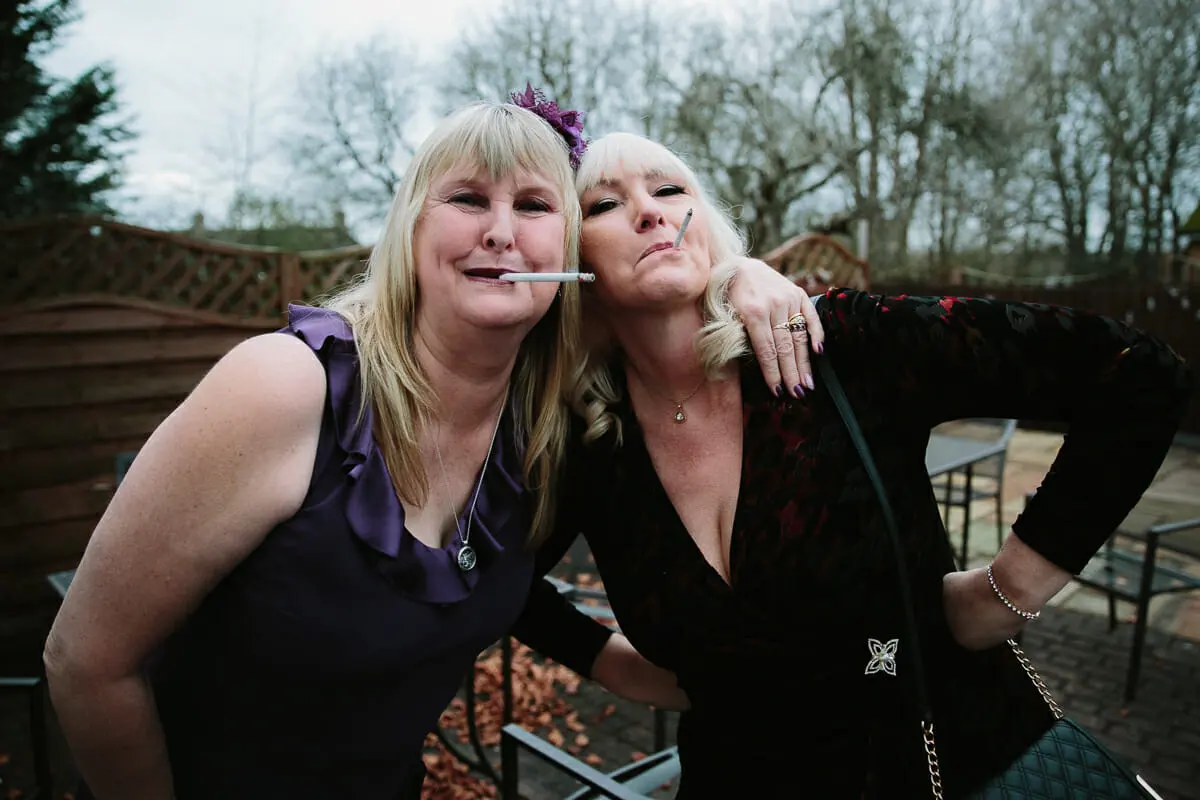 Two women in dark dresses posing arm in arm with candy cigarettes on a patio