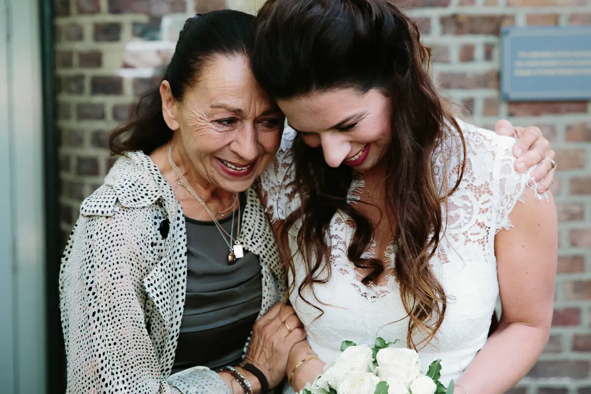 Bride in lace laughing with an older woman as they huddle together by a brick wall