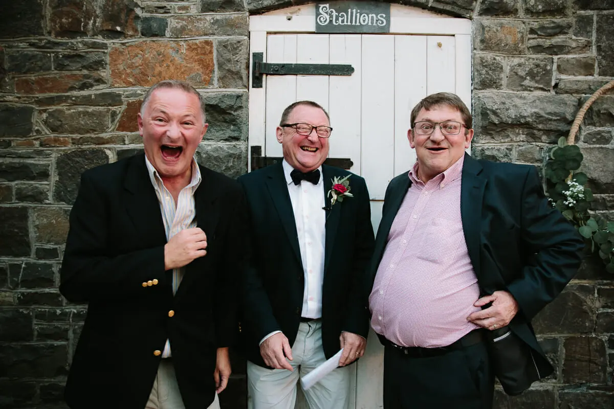 Three laughing men in jackets standing outside a stone stable door