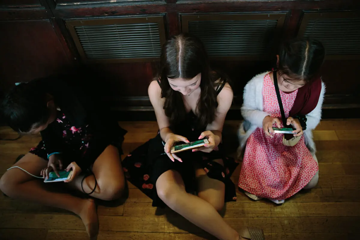 Three young girls sitting on a wooden floor playing handheld games at a wedding