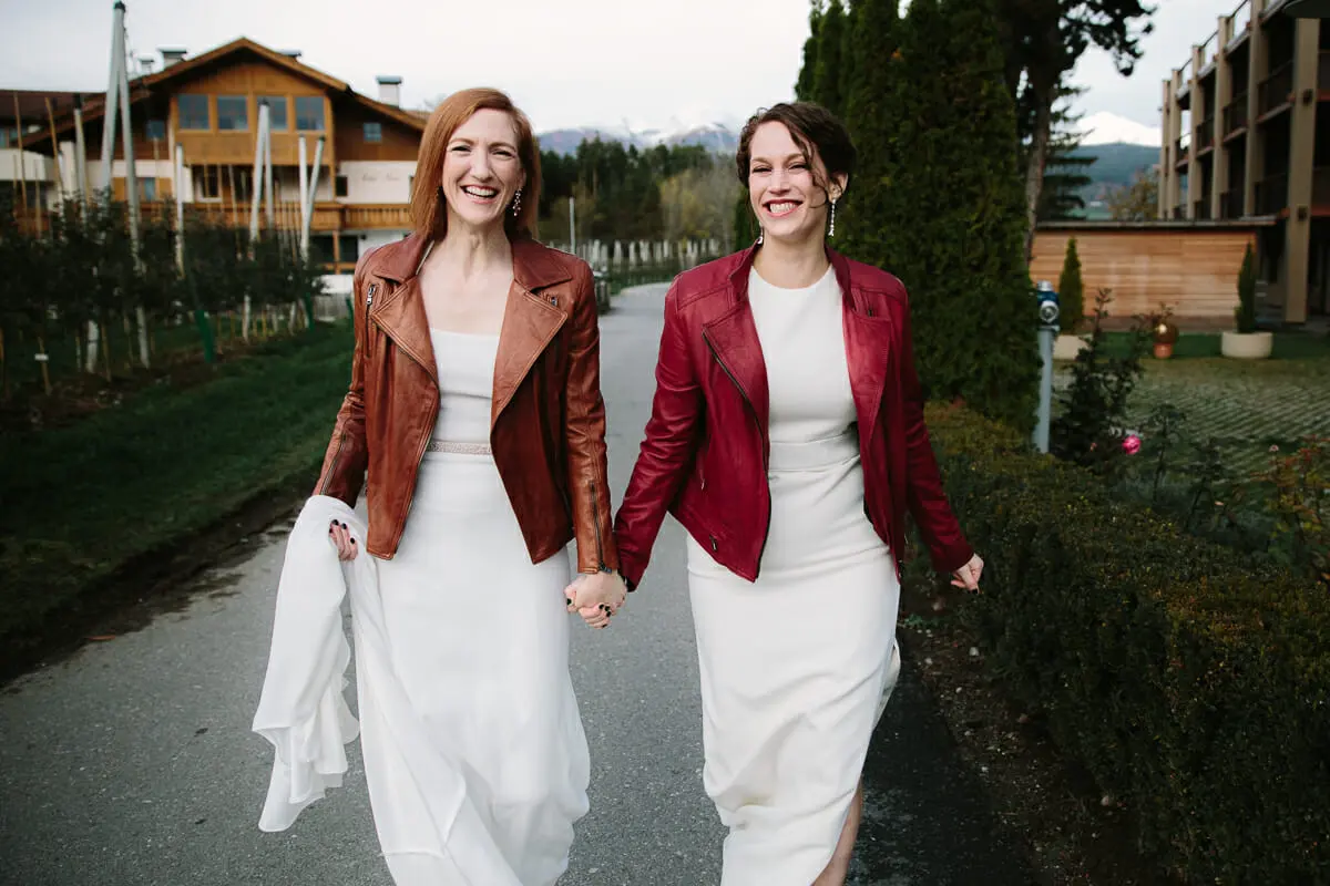 Two brides in white dresses and leather jackets walking hand in hand along a path with mountains behind