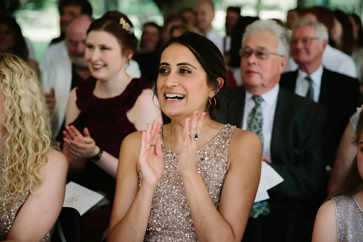 Bridesmaid in a sequinned dress beaming and clapping among seated guests