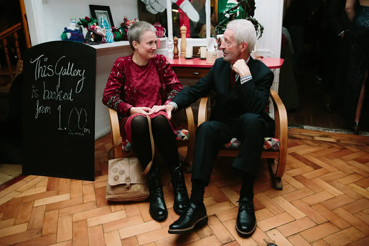 Older couple chatting in wooden chairs on a parquet floor beside a chalkboard sign at the Jam Factory