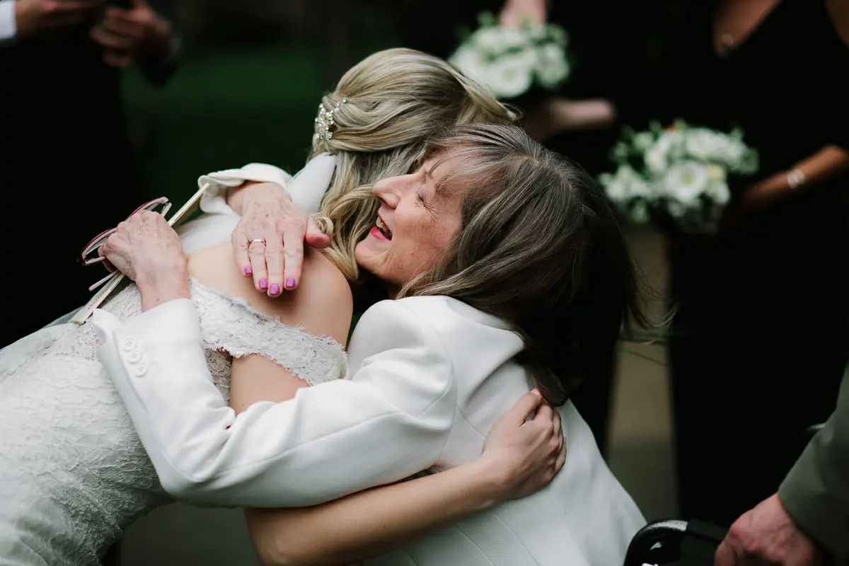 Bride being hugged tightly by a laughing older woman in a white jacket
