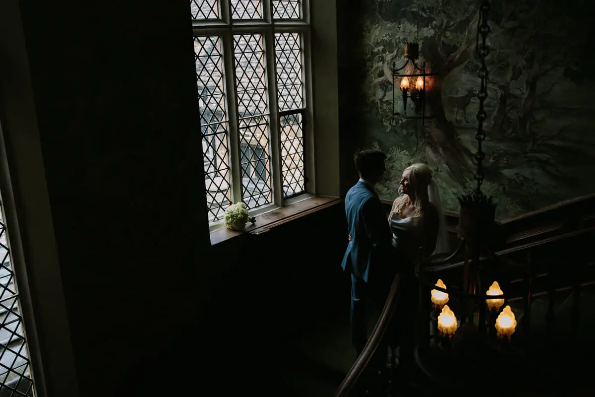 Bride and groom by a leaded window on a dimly lit staircase landing