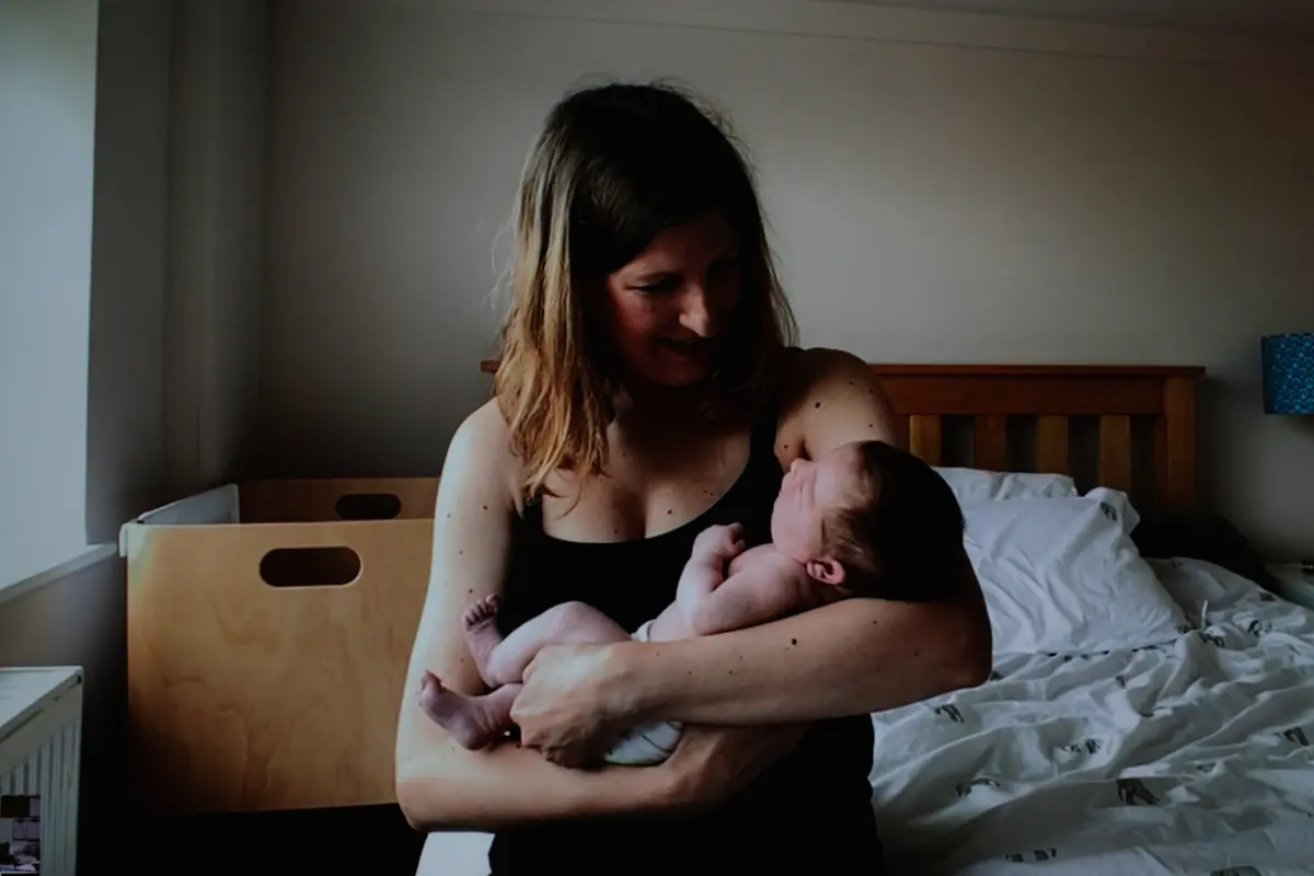Mother smiling down at her newborn baby in her arms beside a bed and wooden crib
