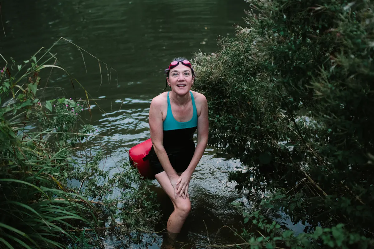 Smiling woman in a swimsuit and goggles wading out of a river with a red tow float