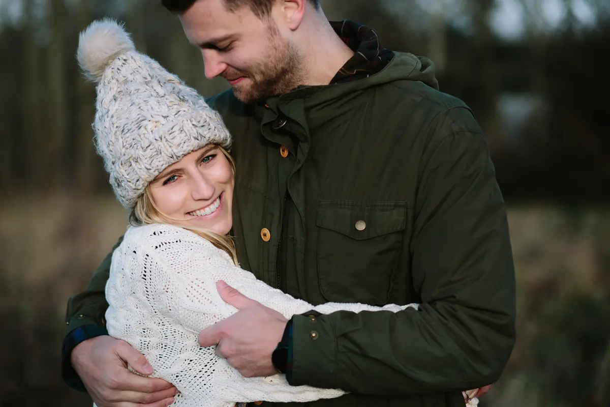 Smiling woman in a bobble hat and cream jumper wrapped in a hug by a man in a green coat