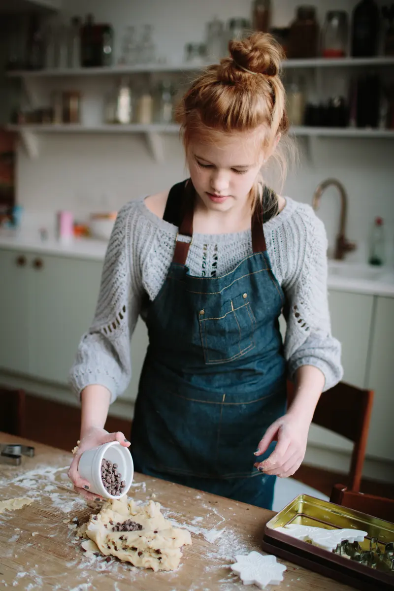 Girl in a denim apron adding chocolate chips to cookie dough on a floury kitchen table