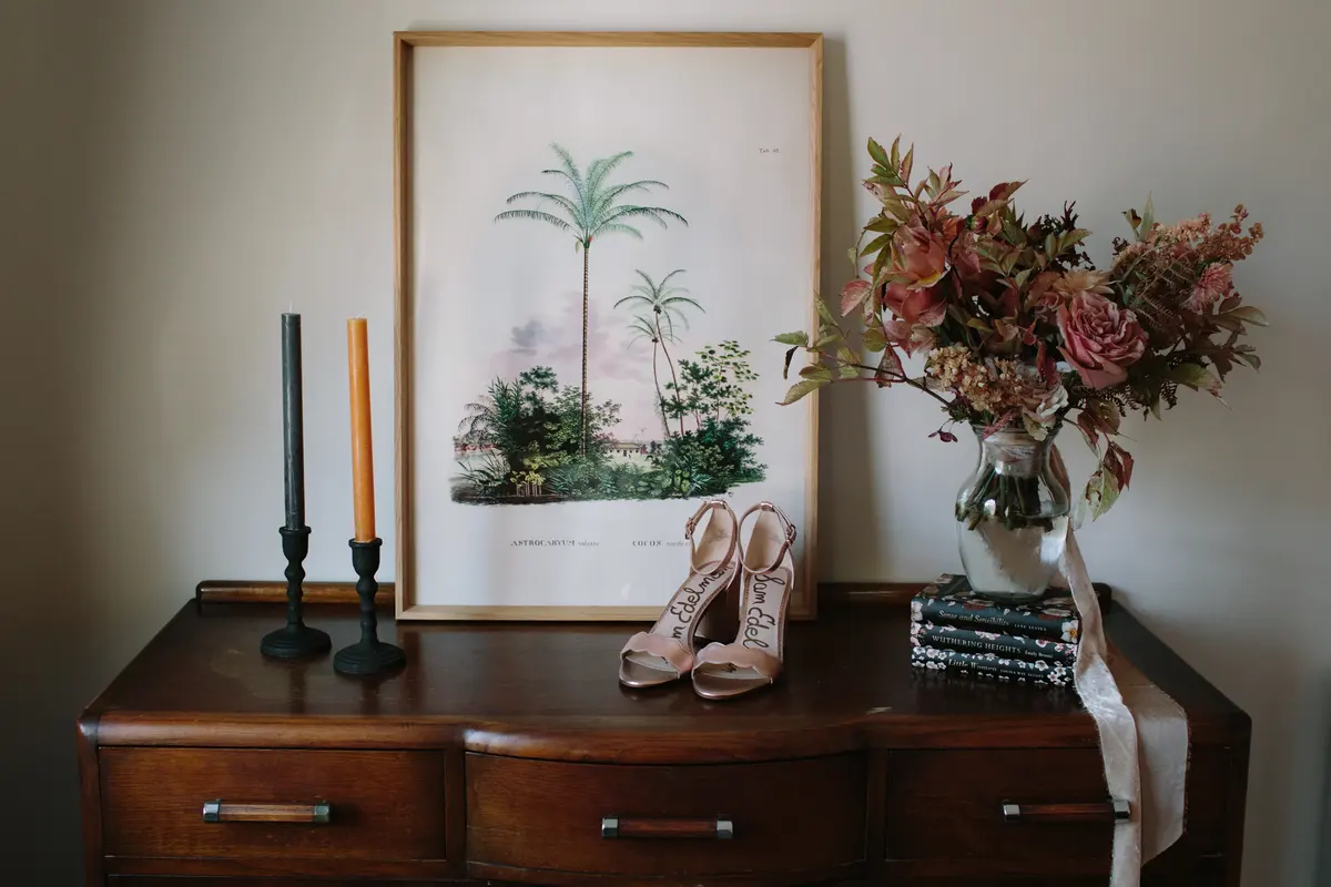 Bridal shoes on a wooden chest of drawers beside a palm print, candles and autumn flowers