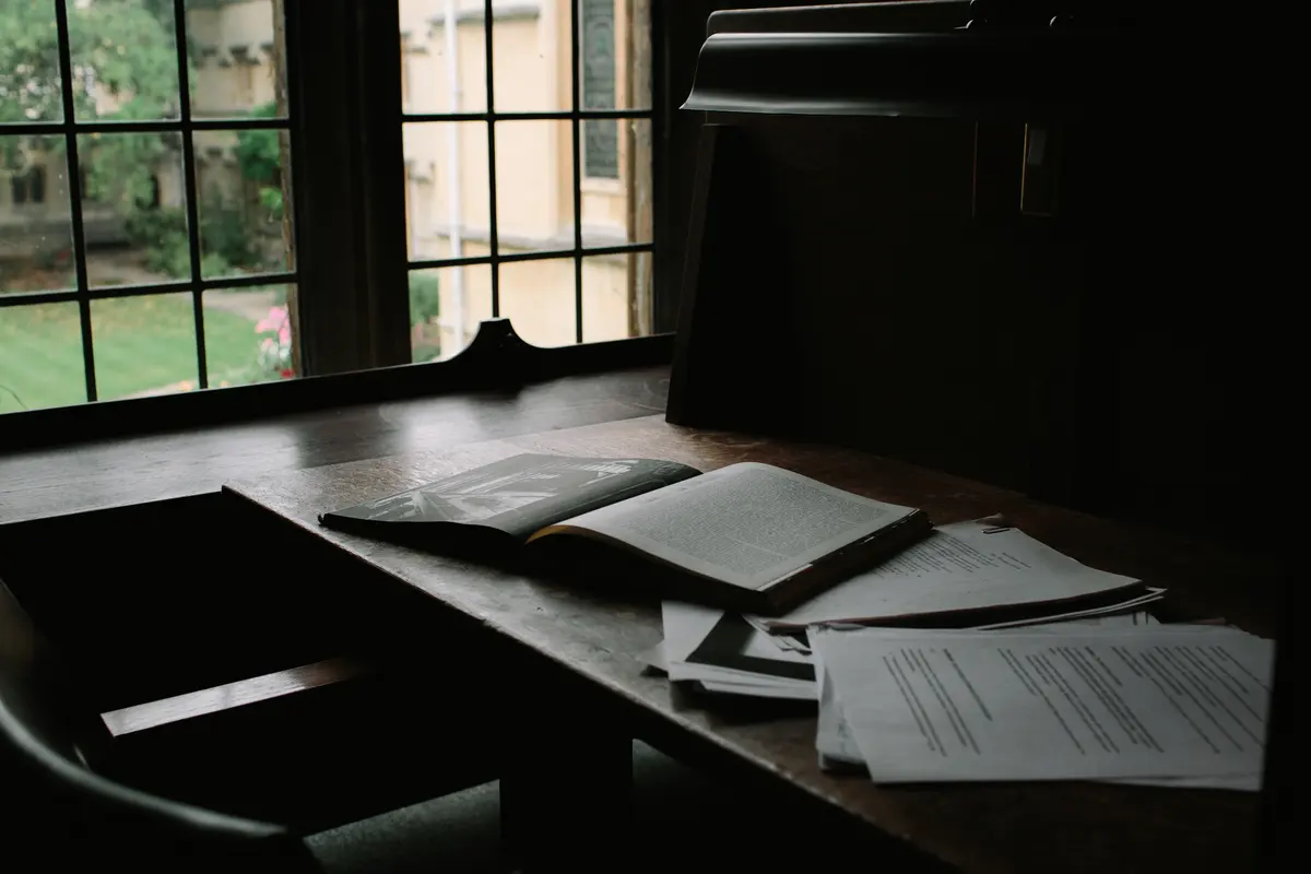 Open book and papers on a wooden desk by a leaded window overlooking an Oxford college lawn