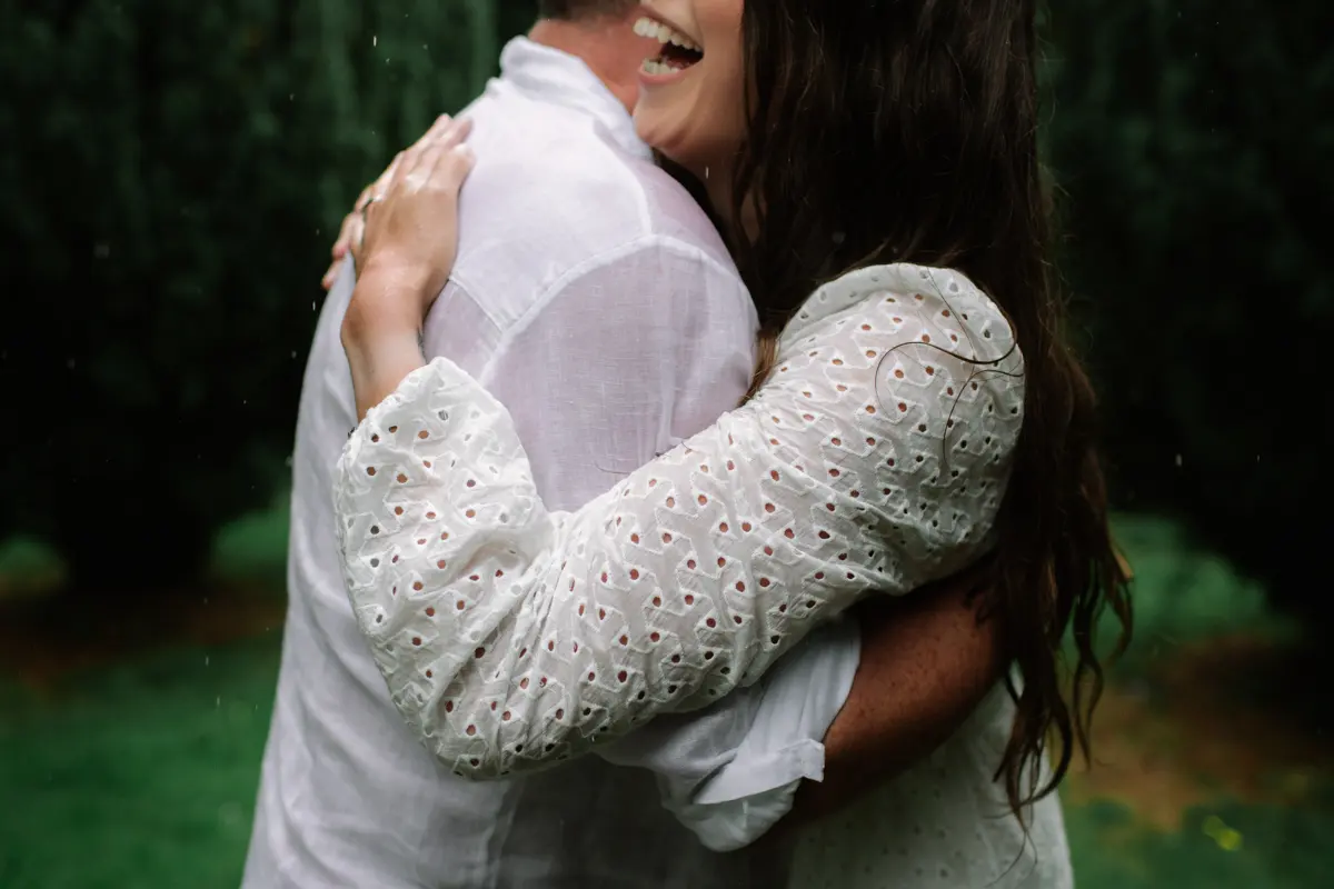 Laughing woman in a white broderie dress hugging a man in a white shirt in the rain