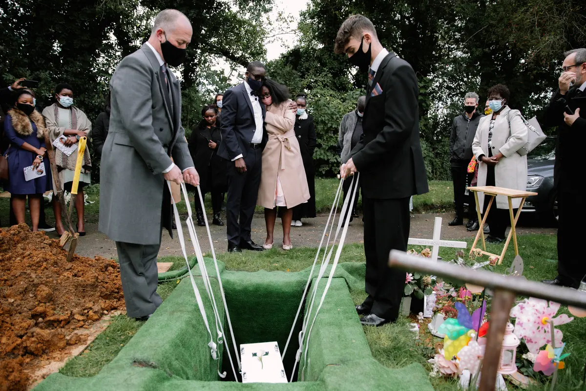 Two men in suits and face masks lowering a small coffin into a grave as masked mourners look on