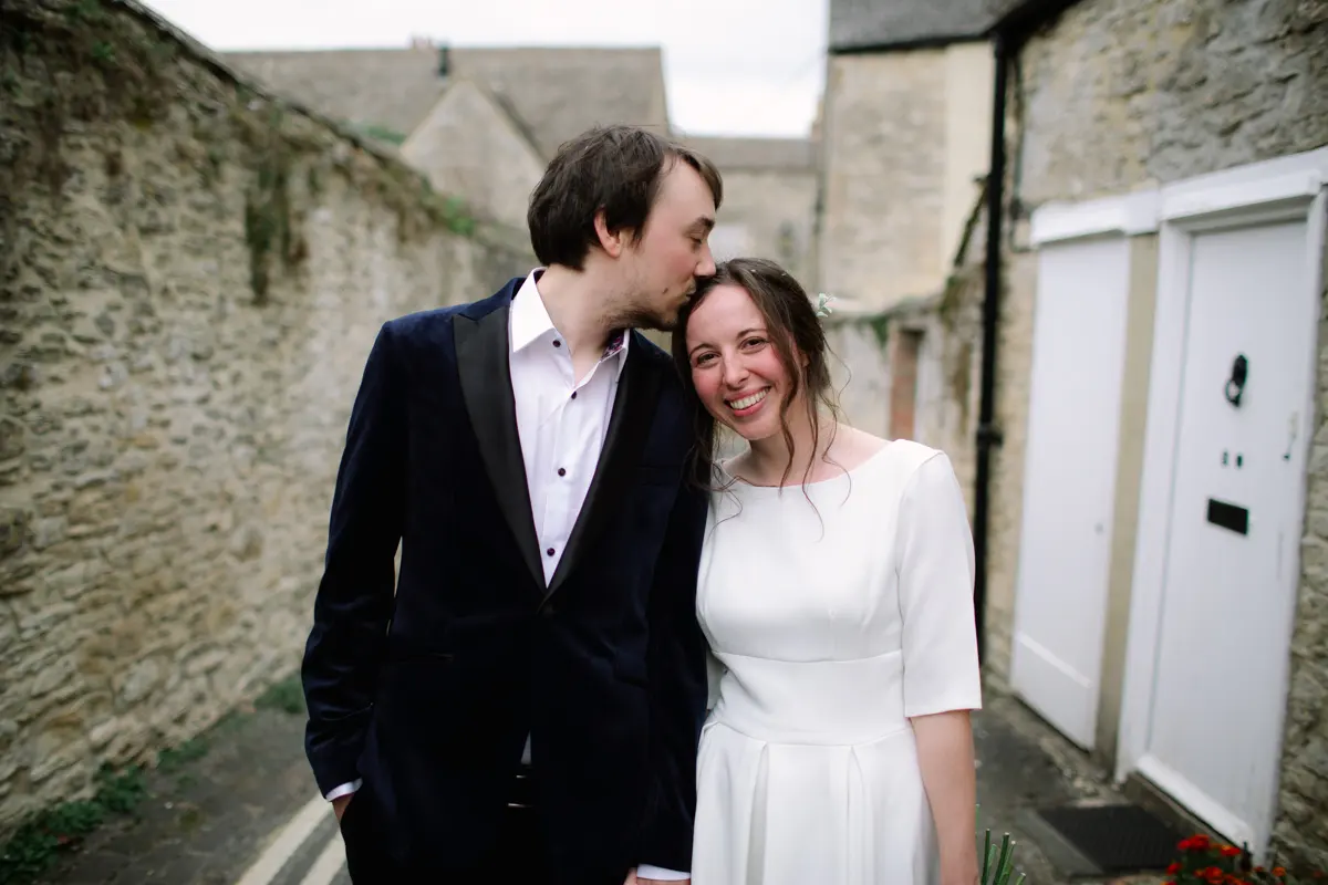 Groom in a velvet jacket kissing the head of his smiling bride in a narrow Cotswold stone lane