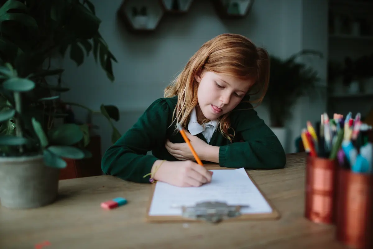 Girl in a green school jumper writing with a pencil at a wooden table beside pots of pens