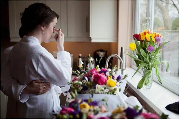 Bride in a white robe wiping her eye as she is hugged from behind beside fresh flowers in a kitchen