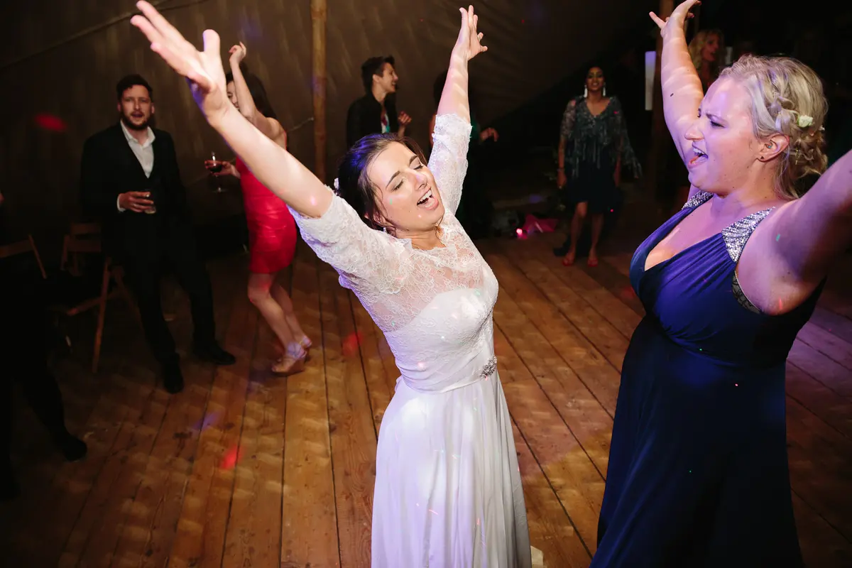 Bride with arms flung up dancing beside a bridesmaid in navy on a wooden dance floor