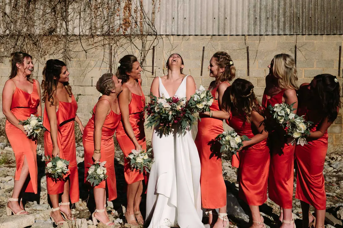 Bride laughing among bridesmaids in orange satin dresses holding white bouquets