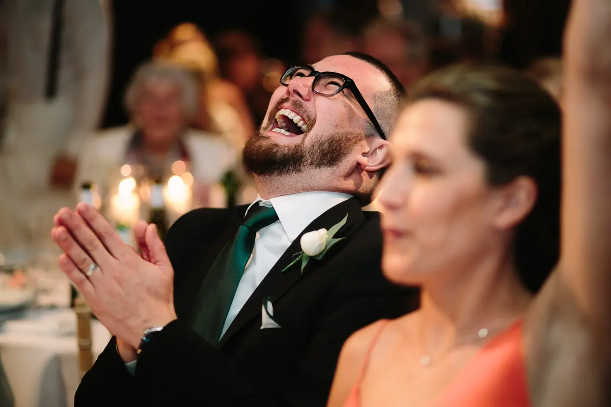 Guest in glasses and a green tie throwing his head back laughing and clapping at the speeches