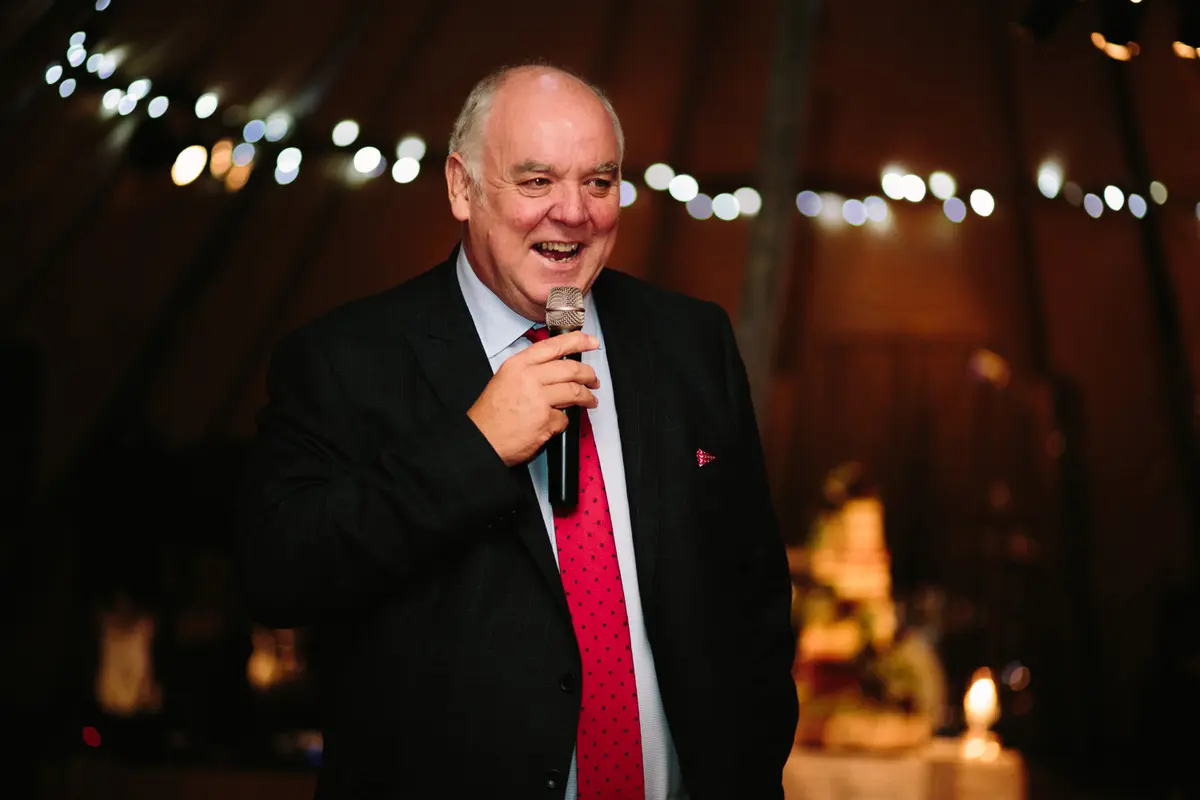 Older man in a red spotted tie smiling as he gives a speech in a fairy-lit tipi