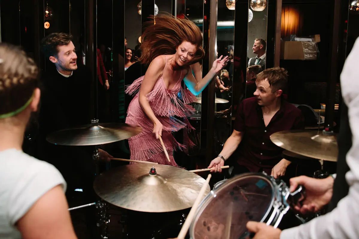 Guest in a pink fringed dress leaping as a drummer and band play in a mirrored bar