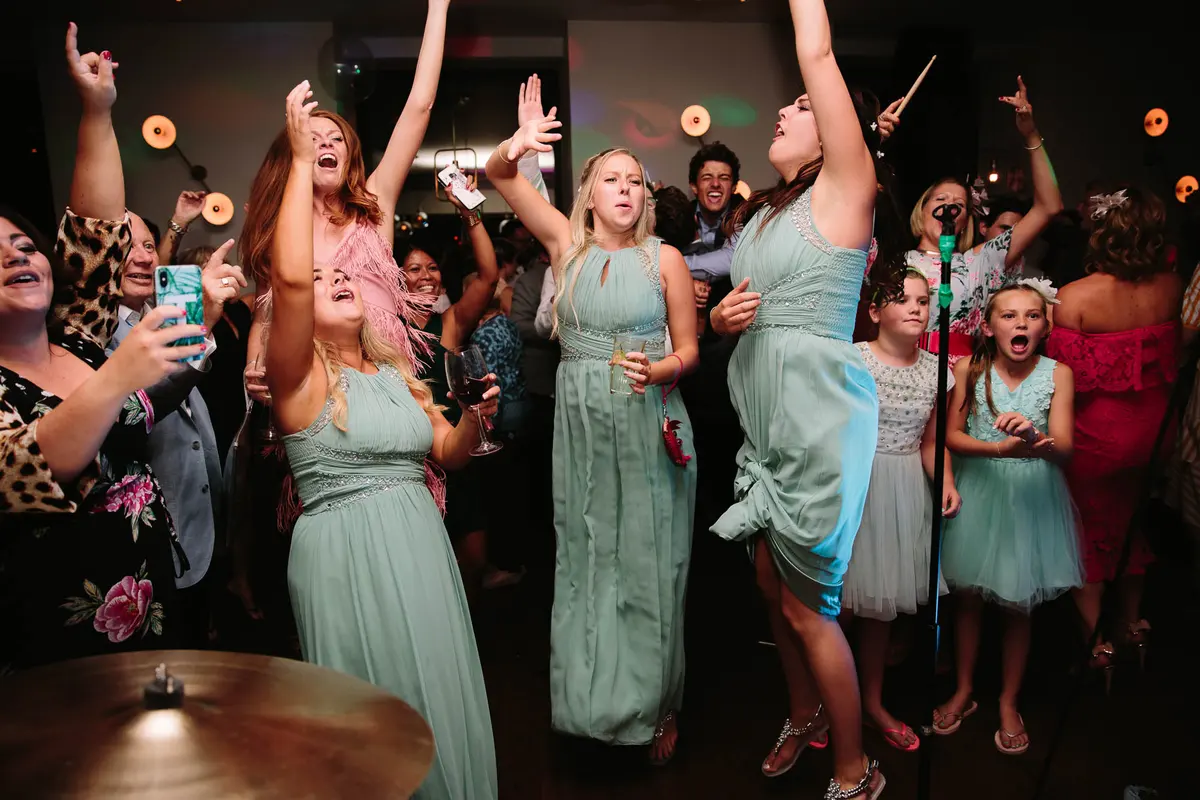 Bridesmaids in sage green dresses jumping and cheering with arms raised on the dance floor