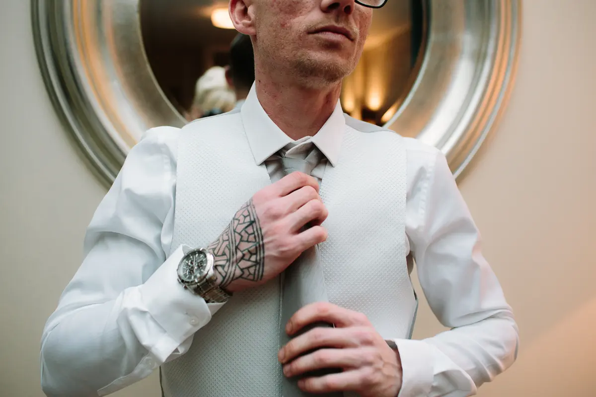 Groom with a tattooed wrist straightening his tie in front of a round mirror