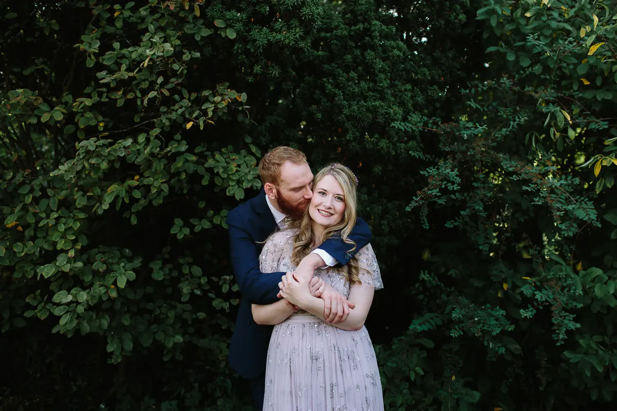 Groom hugging the bride from behind in front of dark green foliage
