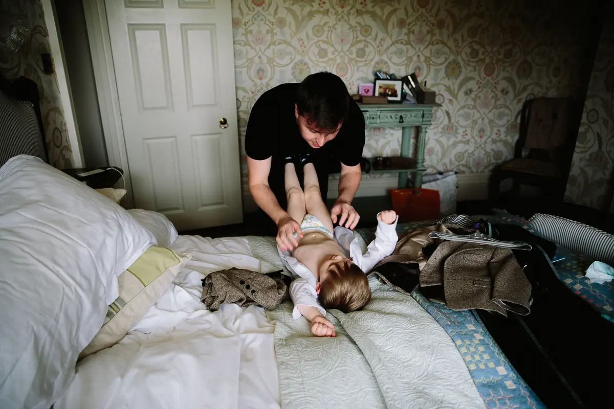 Father dressing a toddler in a tweed suit on a bed before the wedding