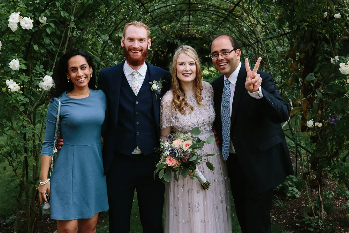 Bride and groom posing with two friends under a rose arch, one making a peace sign
