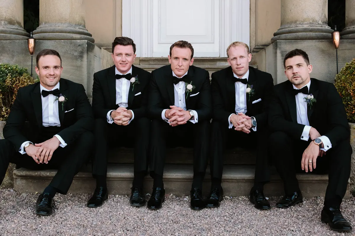 Five groomsmen in black tie sitting on the stone steps of a grand house