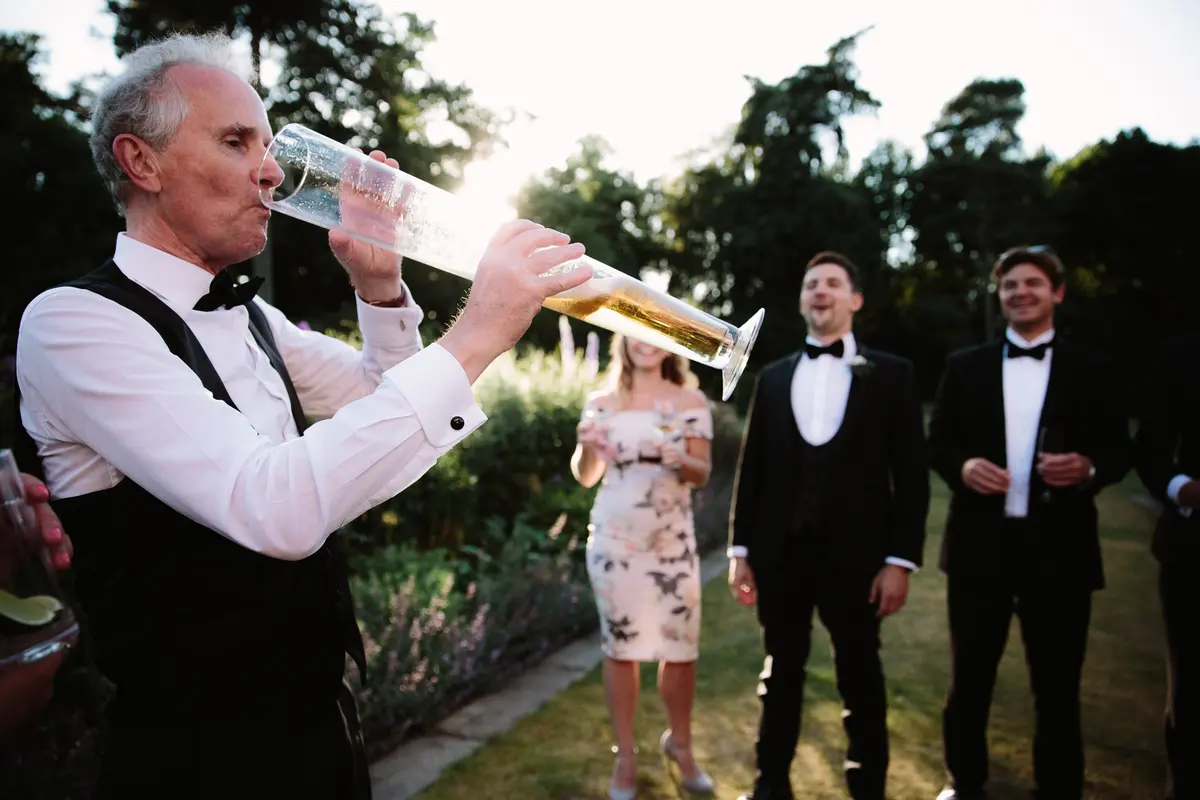 Older man drinking beer from a yard glass as men in black tie laugh in a garden