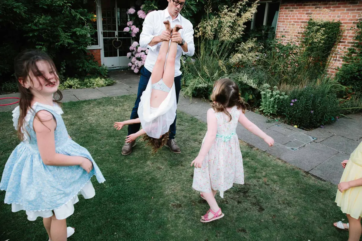 Man holding a girl upside down by her ankles as other girls in party dresses spin on the lawn
