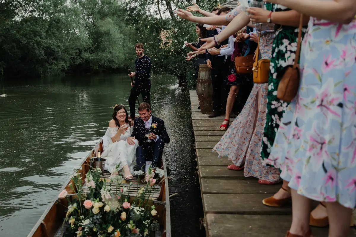 Guests throwing confetti from the jetty as the bride and groom sit in a flower-filled punt at Cherwell Boathouse