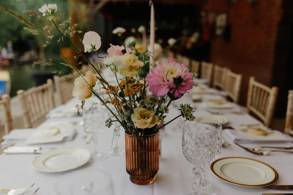 Wedding table laid with dahlias and sweet peas in an amber vase, cut glasses and gold-rimmed plates by the river