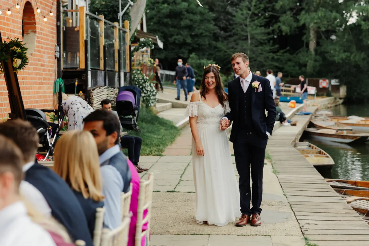 Bride and groom walking hand in hand along the riverside past seated guests at Cherwell Boathouse