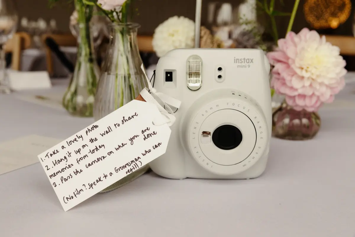 Instax camera on a table with a handwritten tag asking guests to take photos and share memories