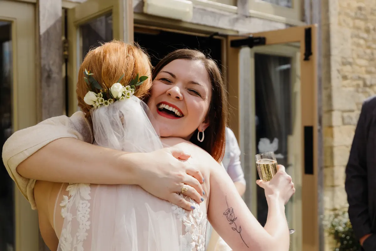 Guest laughing as she hugs the bride, glass of fizz in hand, outside Caswell House